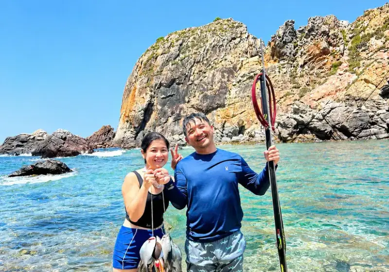 Two guests standing in clear water at Hòn Sẹo with a speargun and a line of fish, rocky headland behind