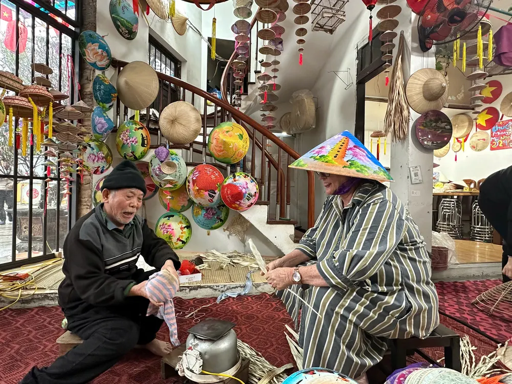 Two artisans in Chuong village stitching palm leaves onto bamboo frames, painted conical hats hanging overhead
