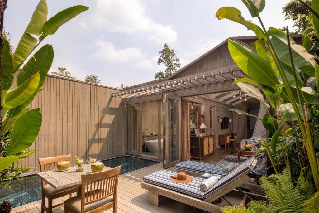 The Garden Pool Villa at An Lam Retreats Saigon River: a timber sundeck with two striped loungers and a table set with fresh coconuts, a dark granite plunge pool along a wooden fence, banana plants, and folding glass doors open into the villa
