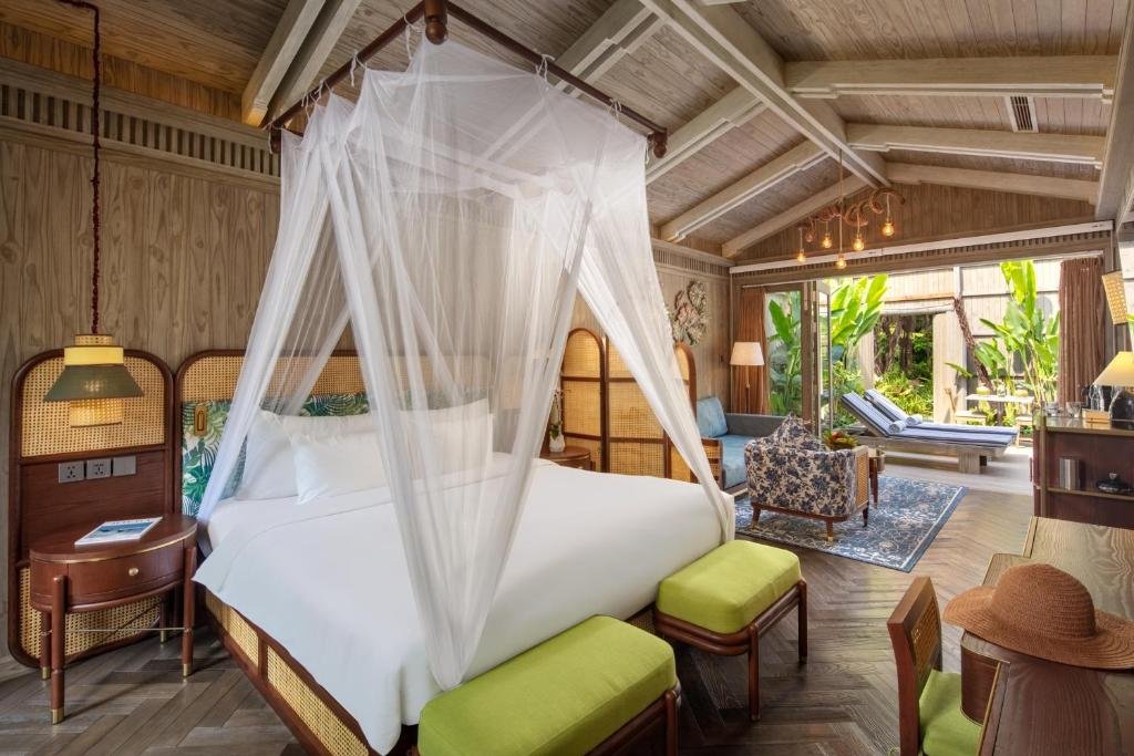 A king bed under a white mosquito-net canopy with rattan headboards and lime-green benches, and beyond it a sitting area with a blue sofa and patterned armchair opening onto the sundeck and garden