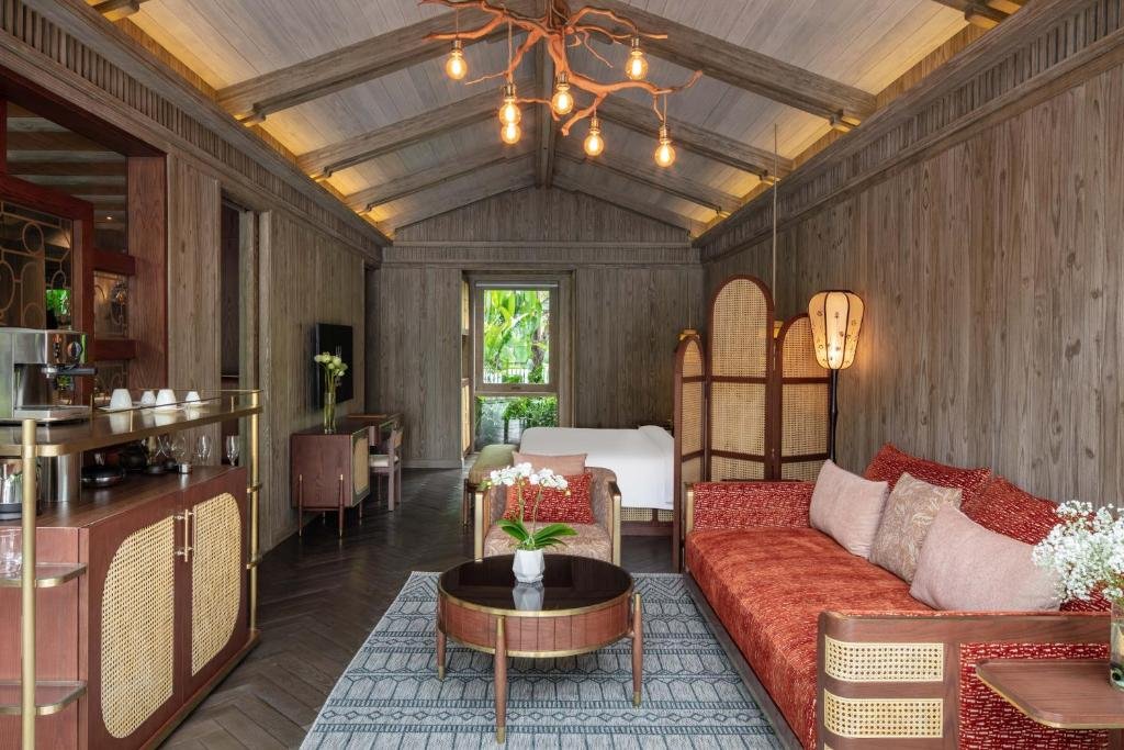 A living area under a vaulted timber ceiling with a branch chandelier: a red cushioned sofa, an armchair and a coffee table with orchids on a patterned rug, a coffee bar on the left, and the bed behind arched rattan screens