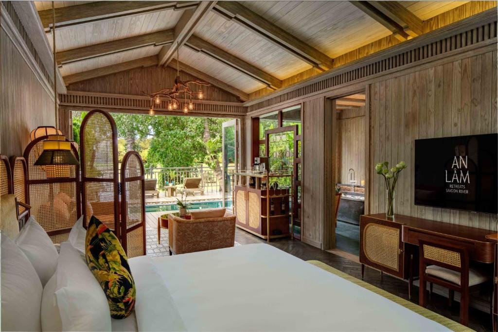 The view from the king bed: rattan screens, an armchair and a drinks cabinet, a desk under a wall-mounted TV, and doors open onto the pool, loungers and trees