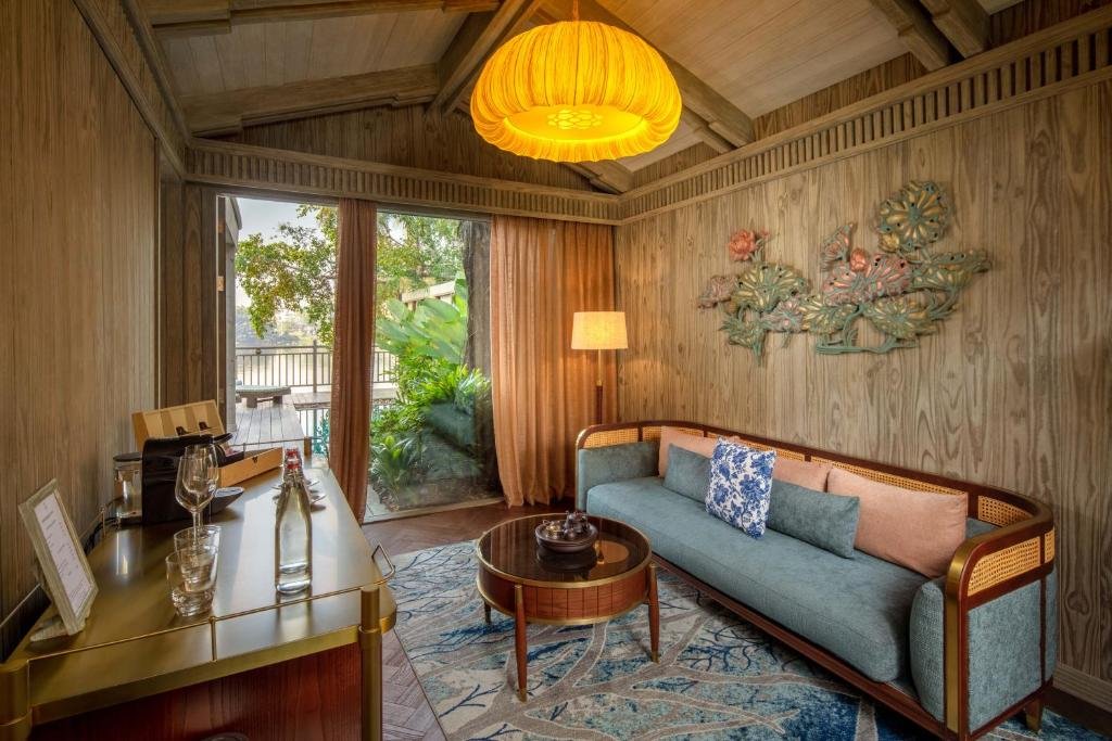 A sitting corner with a blue sofa and a round coffee table under a large silk lamp, a lotus wall sculpture, and glass doors open onto the deck, garden and river