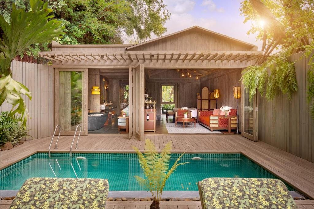 A turquoise private pool with steps and two cushioned loungers on a timber deck, and the villa behind it with its glass doors folded back to the living room, bed and bathtub