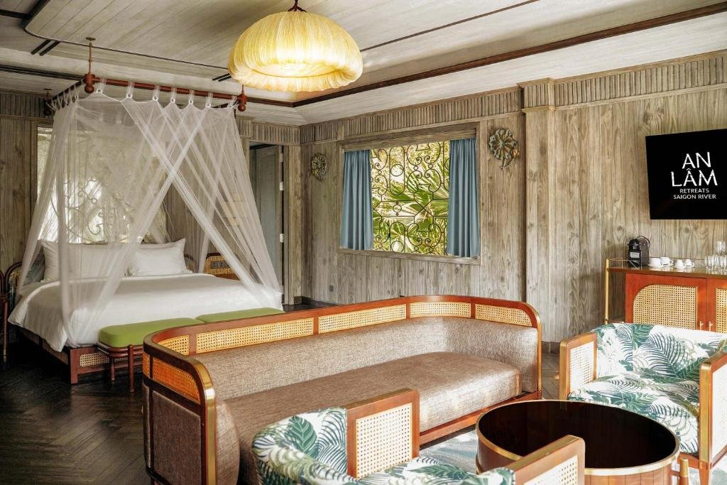 The bedroom: a king bed under a white mosquito-net canopy with a lime-green bench, a rattan-backed sofa and leaf-print armchairs around a round table, a patterned window and a coffee machine on a rattan cabinet