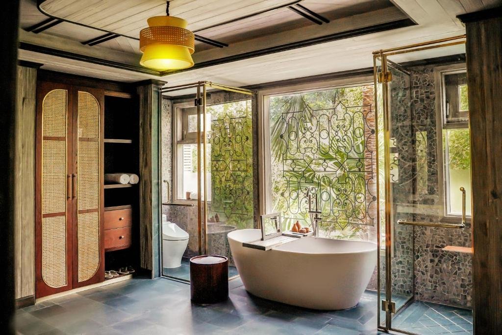 A slate-floored bathroom with a freestanding bathtub before a tall window with wrought-iron tracery, a brass-framed glass shower lined with mosaic stone, a toilet and a rattan wardrobe