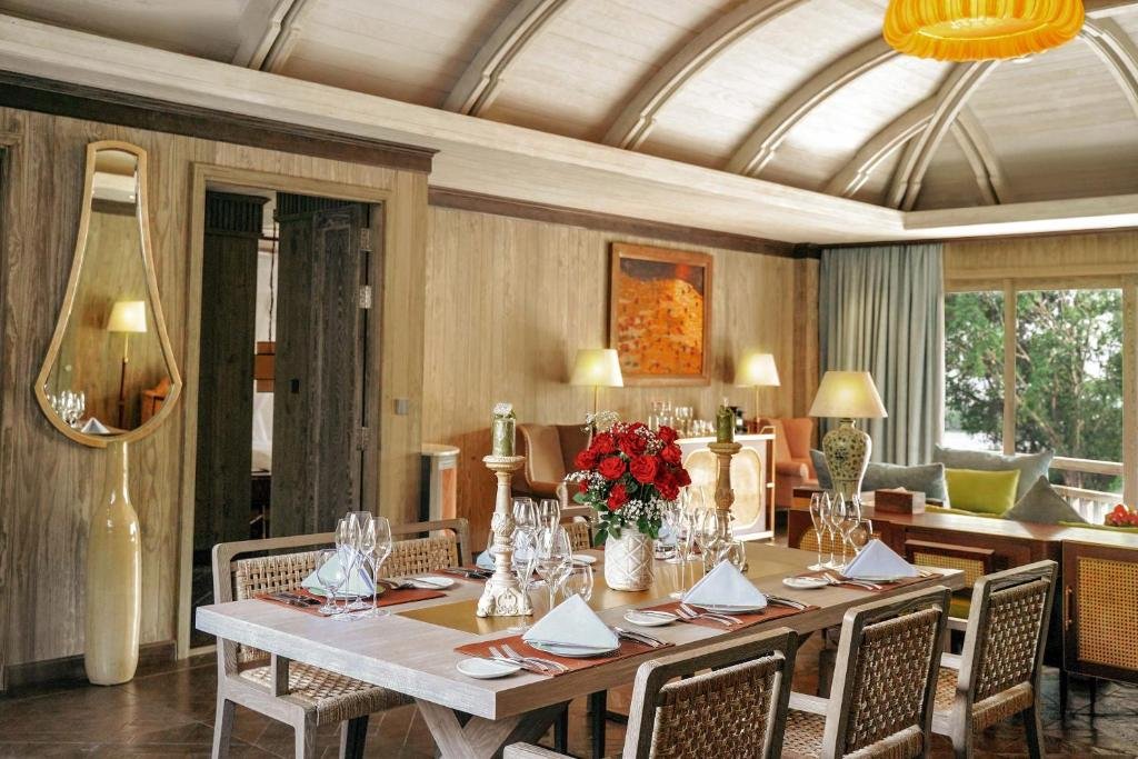 A long wooden dining table with woven chairs, laid for six with folded napkins, wine glasses, candlesticks and red roses, and the lounge and windows beyond