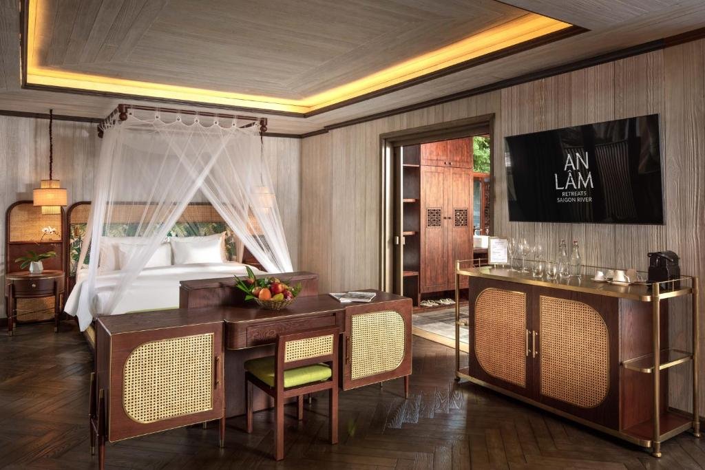 The suite's bedroom: a king bed under a white four-poster canopy with rattan headboards, a rattan-panelled desk with a fruit basket, a drinks cabinet with a coffee machine and glasses, and a wall-mounted TV