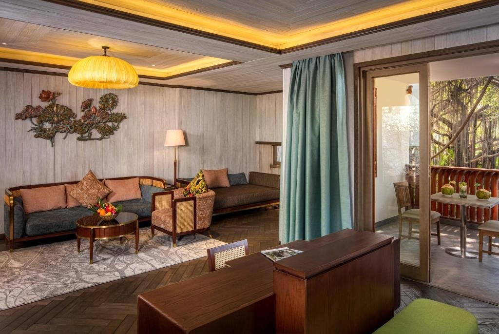 A living room with two cushioned daybeds, a rattan armchair and a round table on a patterned rug under a metal lotus wall sculpture, and glass doors open onto a balcony table with coconuts among banyan trees
