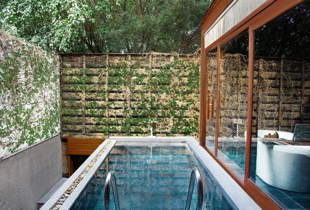 A private plunge pool with steps, walled by timber screens covered in climbing vines under tall trees, beside the glass wall of the bathroom with its bathtub