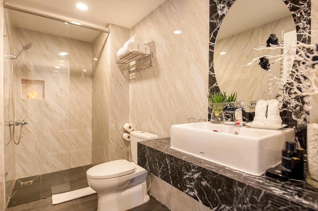 A glass walk-in shower with a hand shower and a lit niche, a towel rack, a toilet, and a white basin on a black marble counter with folded towels and a small plant