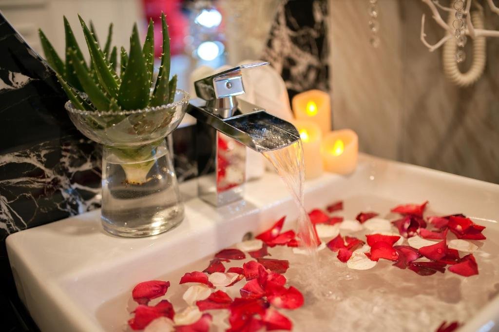 A waterfall tap filling a white basin scattered with red and white rose petals, beside an aloe plant and flickering candles