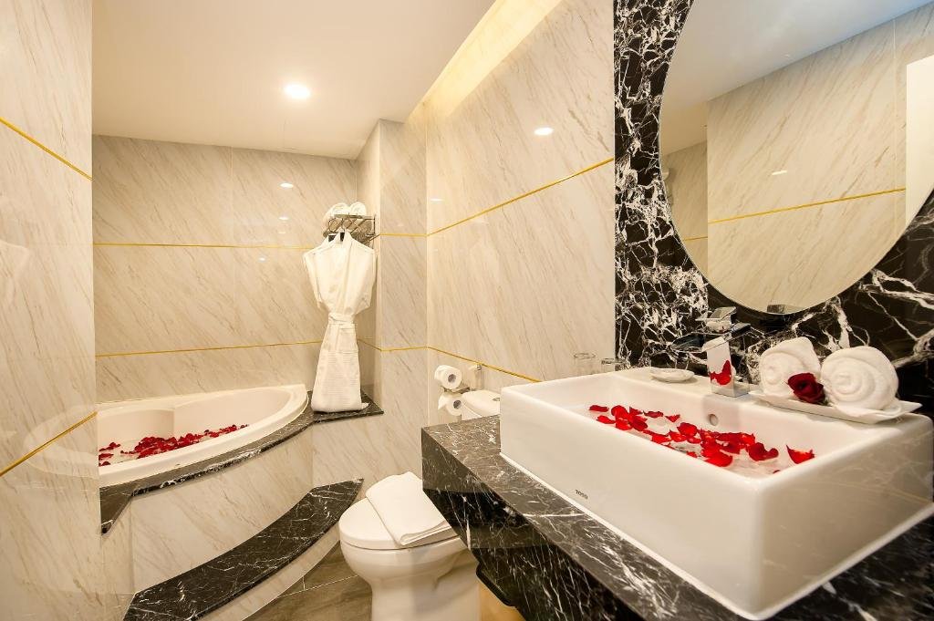 A marble-effect bathroom with gold trim: a corner bathtub scattered with rose petals, a bathrobe on a hook, a toilet, and a rectangular basin on black marble under a round mirror