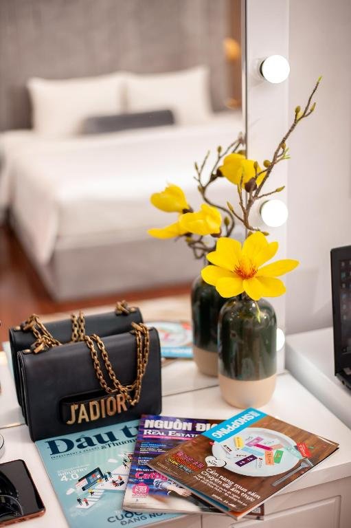 A white vanity with bulb lights, yellow flowers in green vases, a handbag and magazines, and the bed out of focus behind