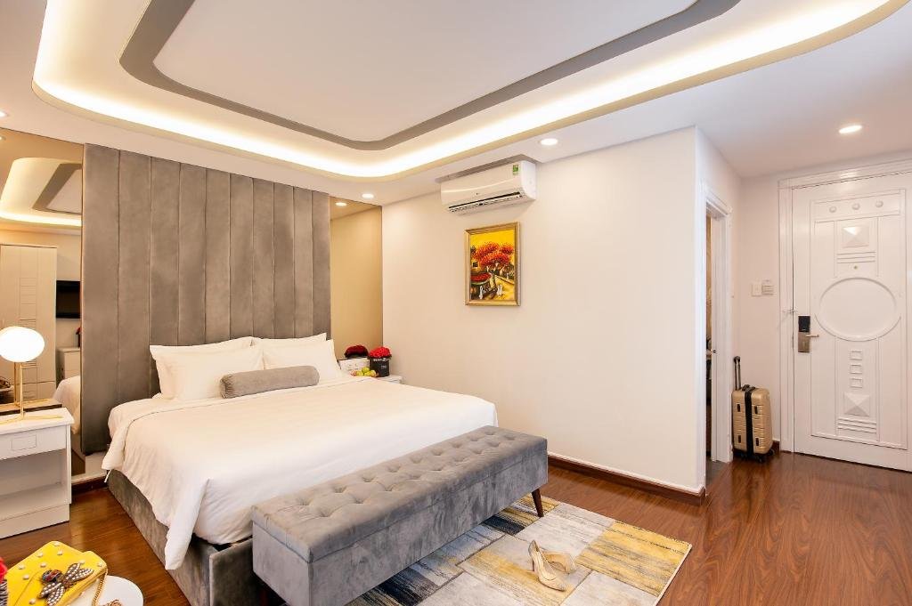 The bed and tufted bench on a patterned rug, a wall-mounted air conditioner above a small painting, a suitcase by the carved white entry door, and wood floors
