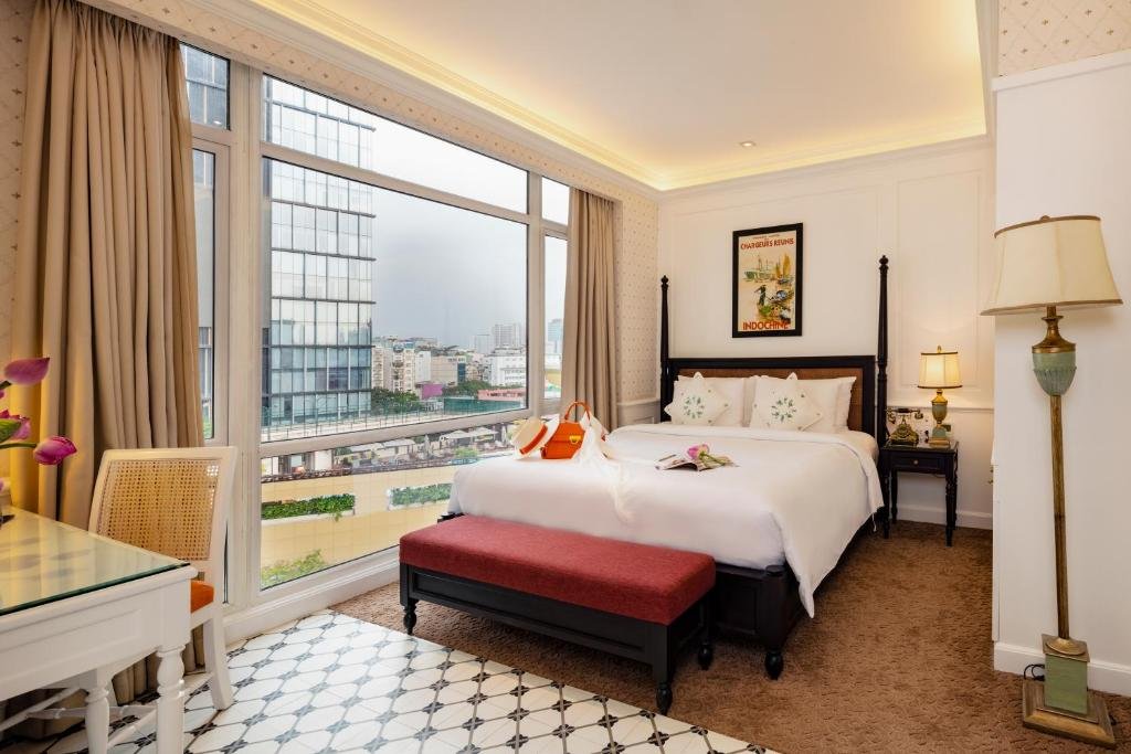 The Vintage Charm Premier Deluxe room at Alagon D&rsquo;antique: a dark four-poster double bed with a red upholstered bench, tall corner windows with beige curtains looking over District 1, a white desk with a rattan chair, a standing lamp and a vintage Indochine poster