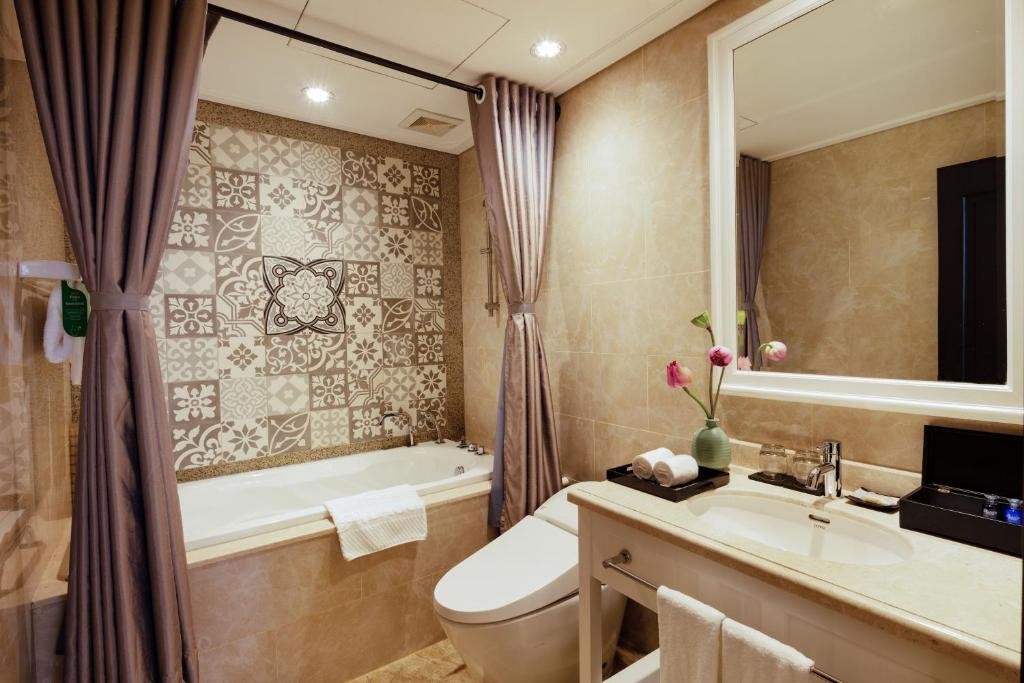 A bathroom with a bathtub under patterned tiles and mauve curtains, a smart toilet, and a white vanity with a lotus flower and rolled towels