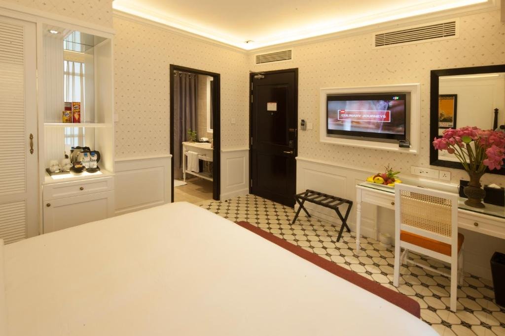 The room from the bed: a white cabinet with a kettle and bottled water, the bathroom and the entry door, a wall-mounted TV over a glass-topped desk with a rattan chair and pink lotus flowers, on patterned cement tiles