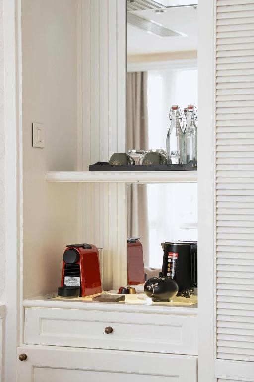 A red capsule coffee machine, a kettle, a tea caddy and glass bottles on white shelves beside louvred wardrobe doors