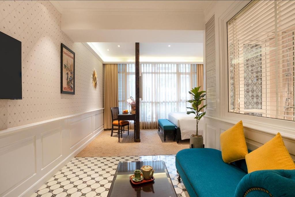 The Indochine Heritage Suite at Alagon D&rsquo;antique: a teal chaise with yellow cushions and a coffee table with a tea set on patterned tiles, a TV and a vintage poster on the wall, and beyond a carved post the bed, a teal bench and a desk before tall curtained windows