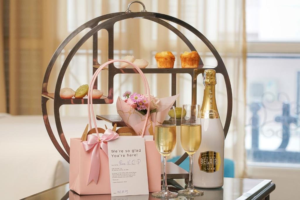 A round metal stand of small cakes and muffins, a pink gift bag with a welcome card, flowers, two glasses of sparkling wine and a bottle, on a glass table by the window