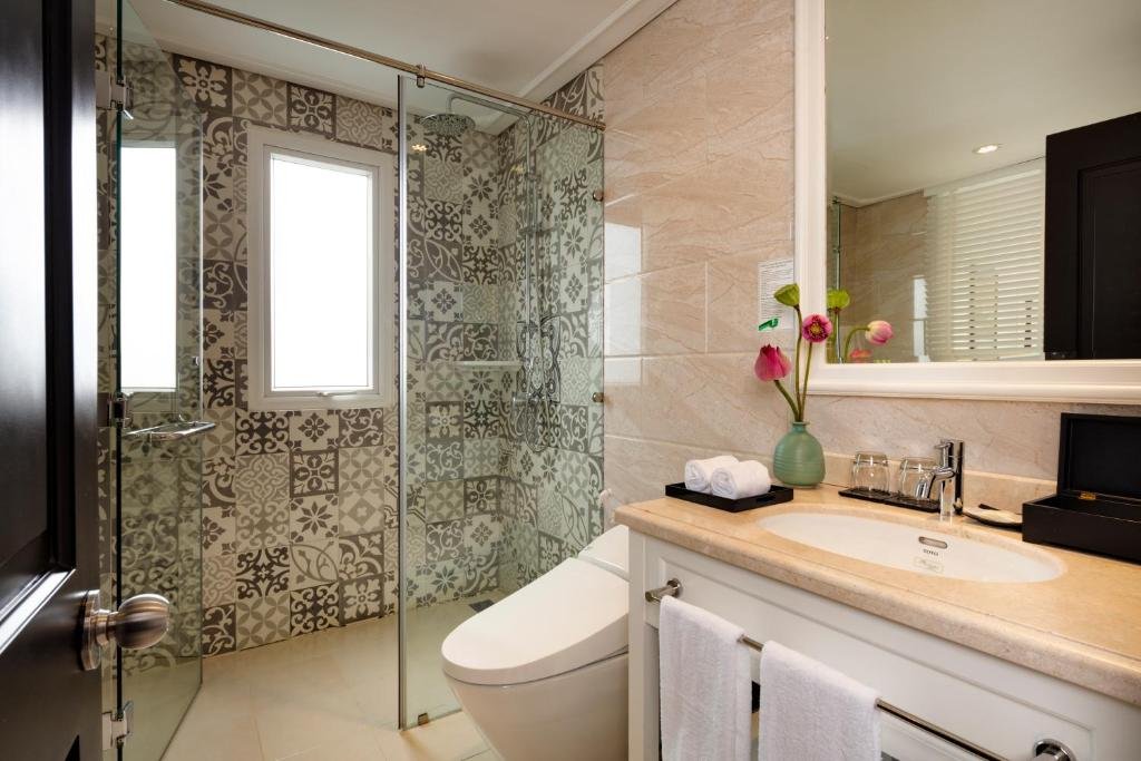 A bathroom with a glass walk-in rain shower lined with grey patterned tiles, a small window, a smart toilet and a white vanity with lotus flowers and rolled towels