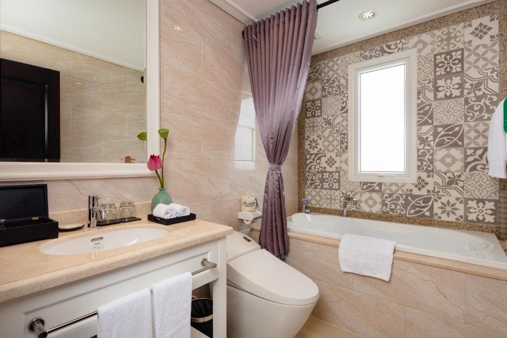 A bathroom with a bathtub under patterned tiles and a small window, a mauve shower curtain, a smart toilet, and a white vanity with a lotus flower and rolled towels