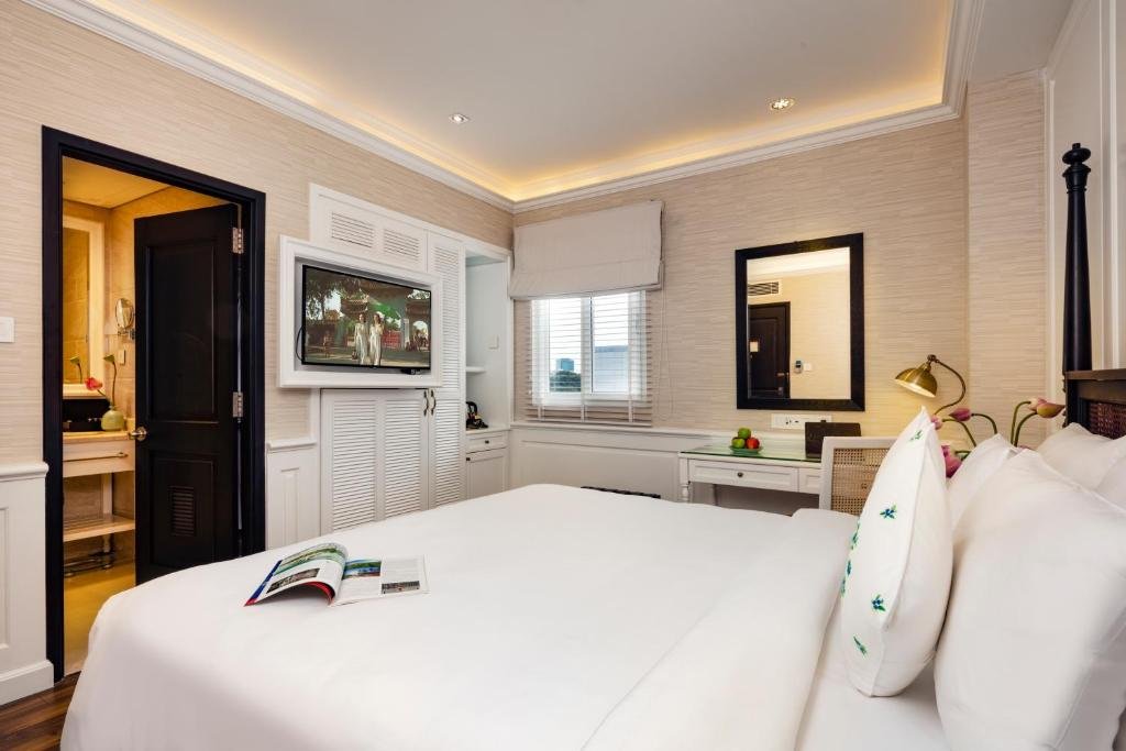 From the bed: a TV set into a white louvred cabinet, the bathroom through a black door, a desk under a black-framed mirror, and a window with a Roman blind