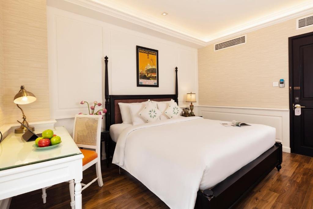 The four-poster bed and the Indochine poster seen past a glass-topped white desk with a bowl of apples, a brass desk lamp and a rattan chair with an orange cushion, on a dark wood floor