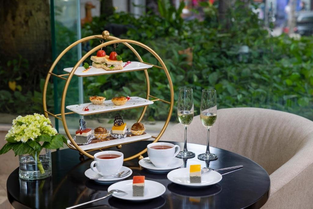 A gold three-tier stand of small sandwiches, tartlets and cakes, two cups of tea, two glasses of sparkling wine and slices of layered cake on a round black table beside a garden window