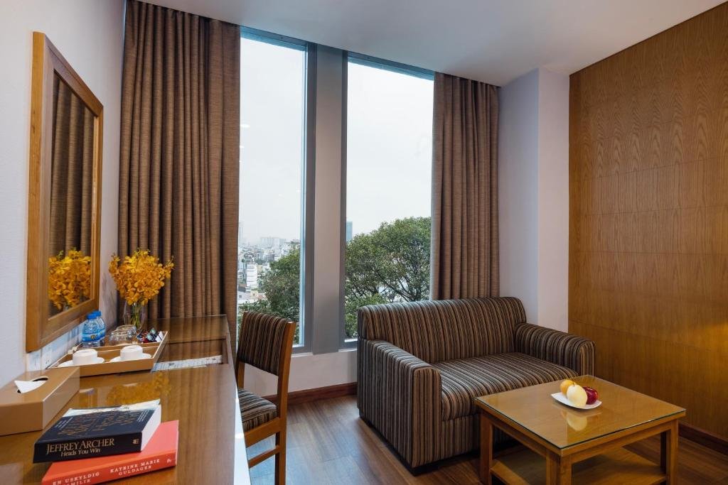 A striped sofa and a glass coffee table with apples beside tall windows onto treetops and the city, and a long wooden desk with orange orchids, a tea tray and books under a mirror