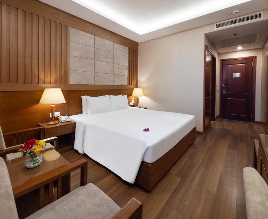 The bed with an orchid on the white cover, bedside lamps and a telephone, a small table with flowers and an armchair in front, and a wood floor leading to the entrance door