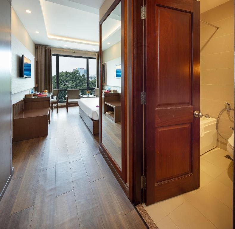 The room seen past the open bathroom door: the bed, desk and TV along a wood floor to the window, a full-length mirror, and the edge of the bathtub on the right