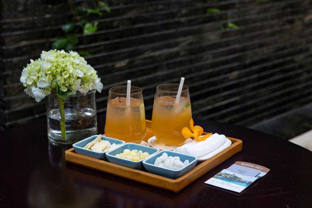 Two iced drinks with straws on a wooden tray with small bowls of dried fruit and lotus seeds, cold towels, an orchid, a vase of hydrangea and a room key folder