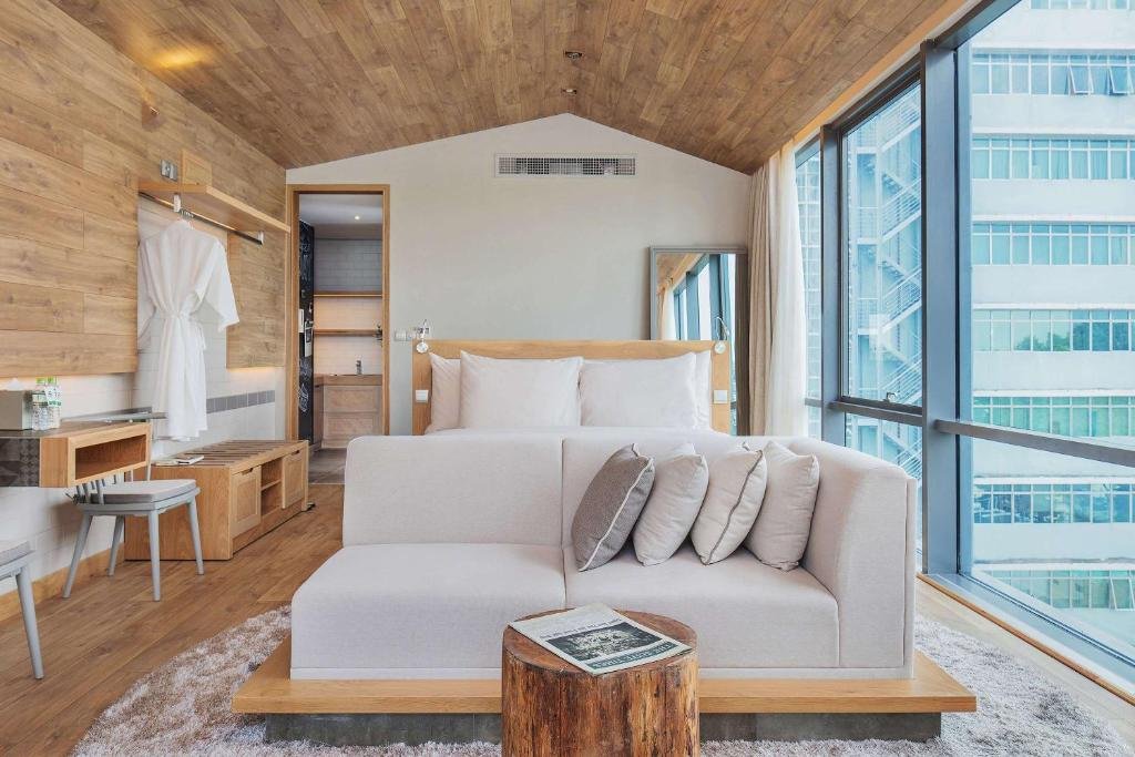 A white sofa with cushions and a tree-stump side table in front of the bed, a bench and a desk under a clothes rail with a bathrobe, the bathroom through a doorway, and a full-height window onto the building next door