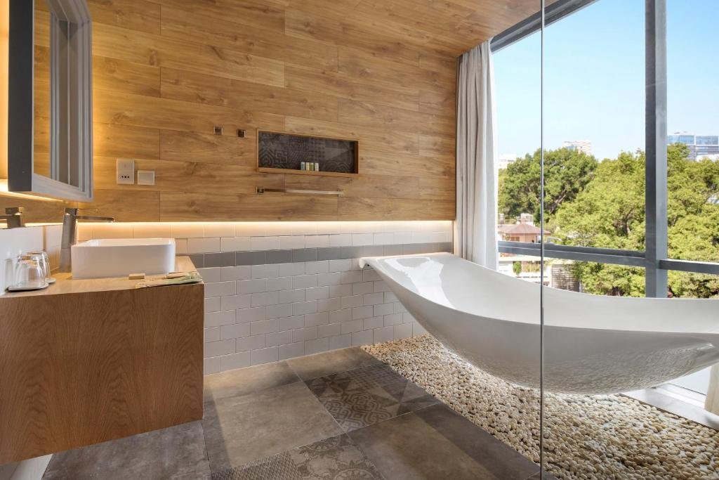 A sculpted white bathtub on a bed of pebbles beside a floor-to-ceiling window onto trees, a basin on a wooden vanity and a timber wall with a toiletry niche