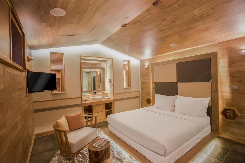 The Double Room without Window at Fusion Suites Saigon: a double bed on a lit platform against a padded checkerboard headboard, a wood-panelled pitched ceiling, a wall-mounted TV, an armchair on a shaggy rug, and an open vanity with a basin and mirrors