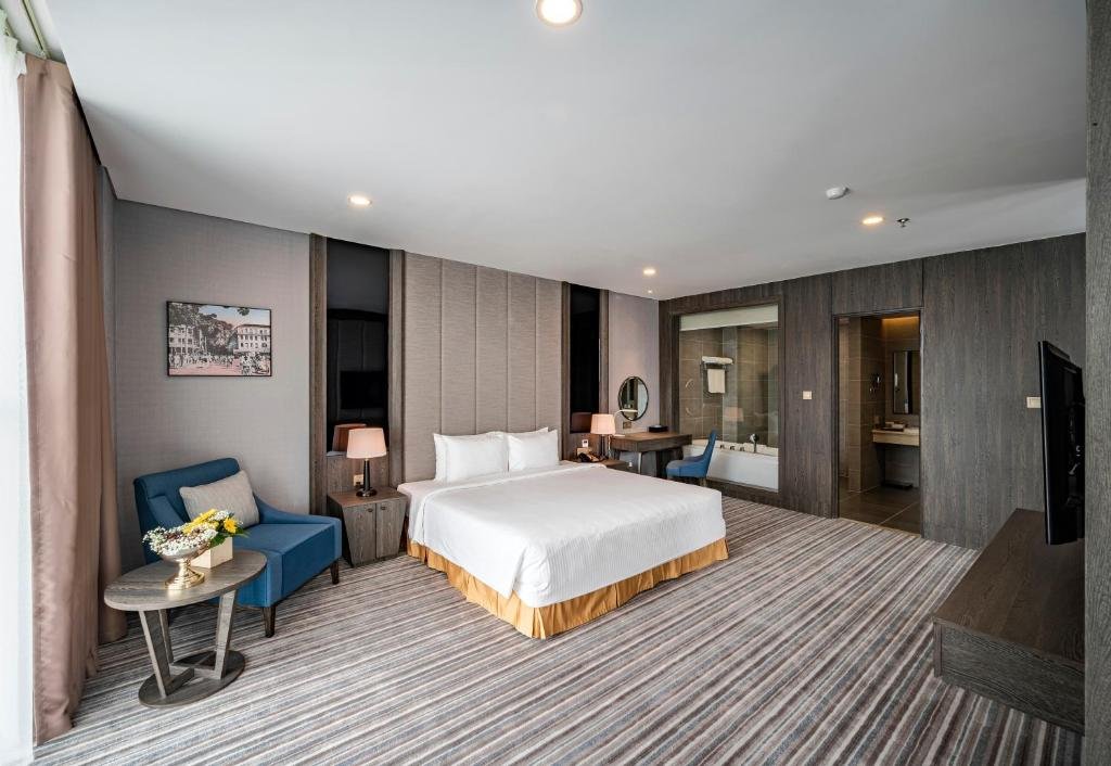 The bedroom: a king bed against a padded grey headboard wall, a blue armchair and side table with flowers, and a glass panel through to the bathtub, with the bathroom door open on the right