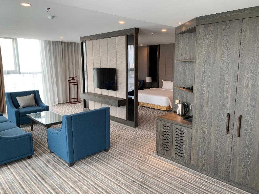 The suite in daylight: blue armchairs and a glass table in front of the TV partition, the bed behind it, and a tall grey wood wardrobe with a kettle on its counter