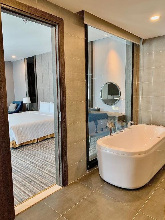 A freestanding white bathtub on grey tiles beside a glass panel, with the bedroom and its king bed seen through the open door
