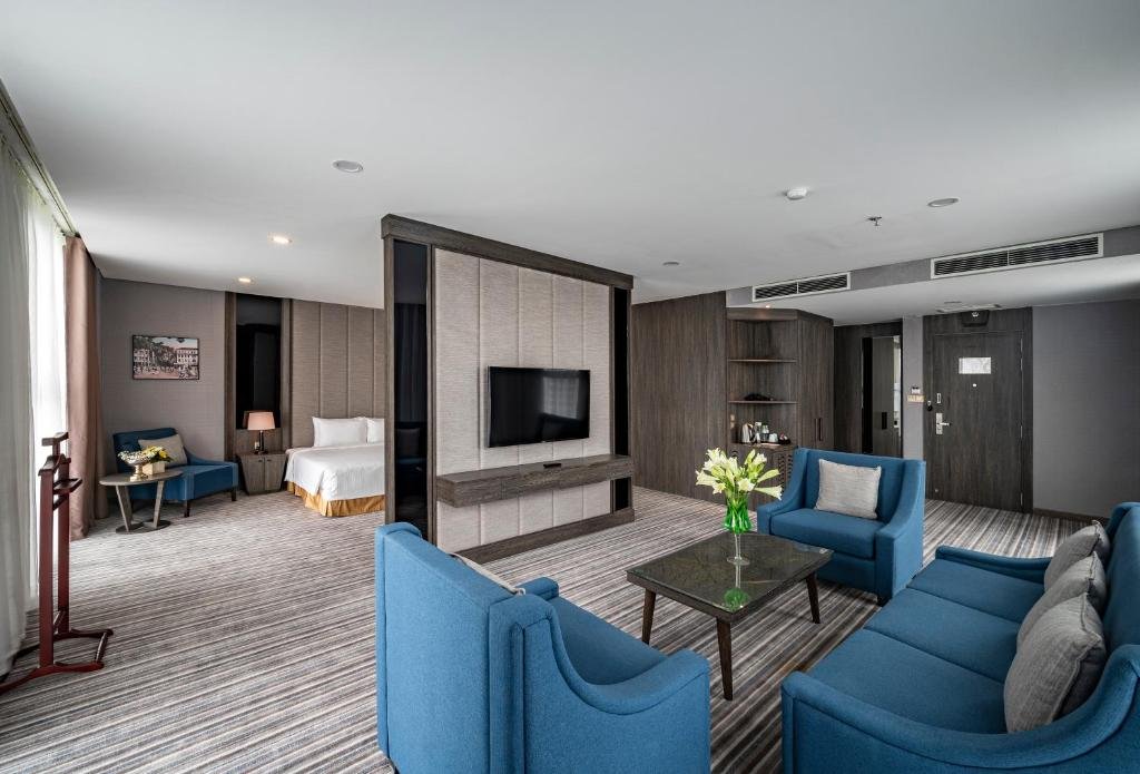 The Suite Executive living room: blue sofa and armchairs around a glass coffee table with white lilies, a TV set into a free-standing partition, and the bed visible beyond it