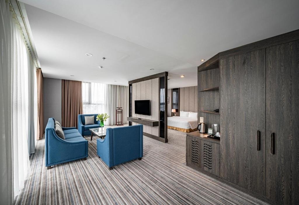 The living room seen from the wardrobe: blue armchairs and sofa by tall curtained windows, the partition with the TV, the bed beyond, and a wood cabinet with a kettle and tea tray