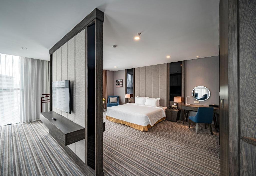 Looking past the TV partition into the bedroom: a king bed with a gold skirt, bedside lamps, a desk with a round mirror and a blue chair, and a window with sheer curtains on the left