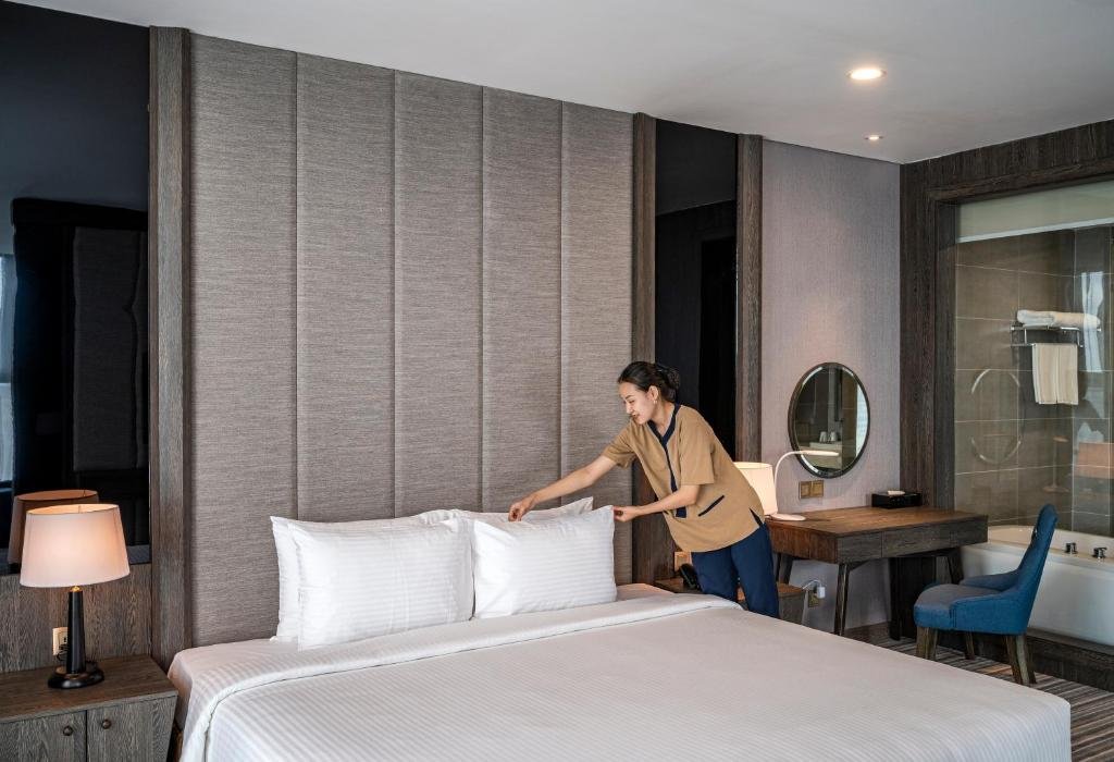 A housekeeper arranging white pillows on the king bed, with the desk, round mirror and the glass view into the bathroom behind