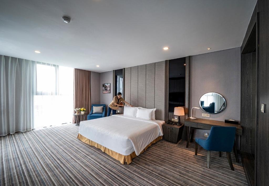The bedroom with a housekeeper smoothing the pillows: a king bed, a blue armchair by the floor-to-ceiling window, and a desk with a round mirror and a blue chair
