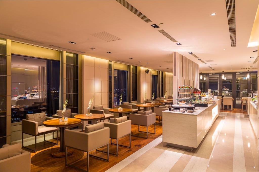 A dining room at night: round wooden tables and upholstered chairs by tall windows, and a long marble buffet counter with a tiered stand of desserts
