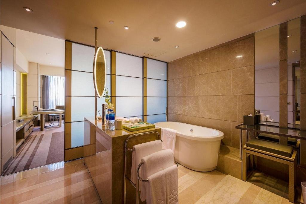 A freestanding white bathtub against marble walls, a towel rail with white towels in front of it, a glass dressing table and stool on the right, and frosted glass panels to the bedroom