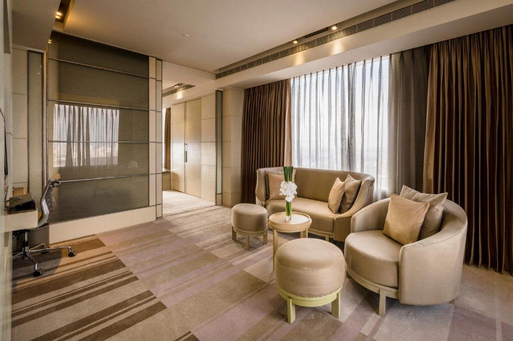 The suite's living room: a curved sofa and armchair with cushions, two round ottomans and a small table with orchids, under a tall window with sheer and bronze curtains, and a desk on the left