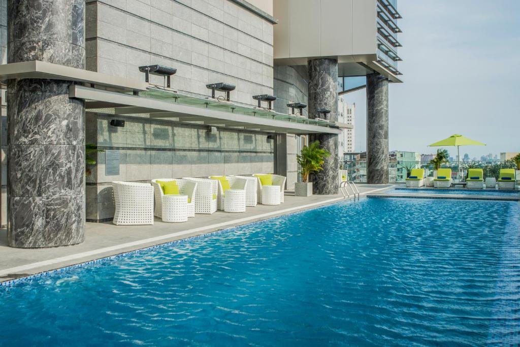A long outdoor pool on a terrace with white wicker seats under marble-clad columns, and lime-green loungers and a parasol at the far end with the city beyond