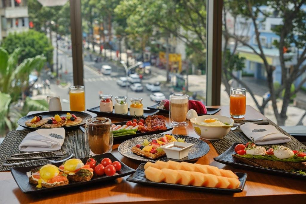 A breakfast table by a window over a tree-lined street: eggs Benedict with smoked salmon, a waffle with fruit, pancakes, sausages and bacon, sliced melon, juices and coffee
